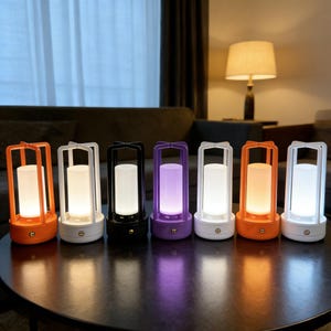 May include: Seven LED lanterns in various colours, including orange, white, black, and purple. Each lantern has a cylindrical light source within a square frame and a round base. The lanterns are displayed on a dark table.