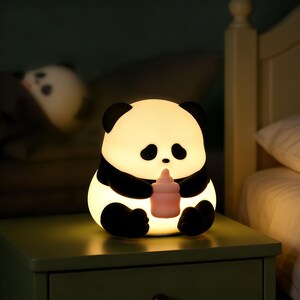 Puede incluir: Luz de noche con forma de panda, con un diseño en blanco y negro, sosteniendo un biberón rosa. La lámpara está sobre una mesita de noche verde. El panda tiene ojos negros y una nariz pequeña.