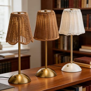 May include: Three table lamps with woven shades in varying colors: tan, brown, and white. Each lamp has a gold-colored base and stem. The lamps are arranged on a wooden surface, with a bookshelf in the background.