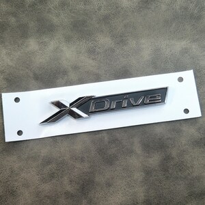 Peut inclure: Un emblème "X Drive" argenté et noir sur un support rectangulaire blanc. L'emblème présente un "X" stylisé et le mot "Drive" dans un rectangle gris foncé. Le support comporte deux trous à chaque extrémité.