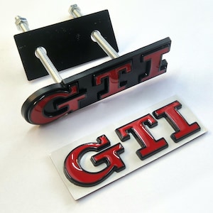 Peut inclure: Un emblème GTI noir et rouge avec deux boulons de montage filetés en métal et une plaque de support noire, accompagné d'un emblème GTI séparé avec lettrage rouge et bordure noire sur une plaque adhésive rectangulaire de couleur argent.