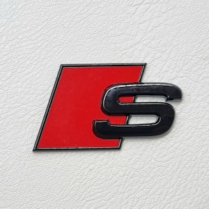 May include: A black and red S emblem, possibly for a vehicle, is set against a textured white backdrop. The emblem has a red trapezoid shape with a black border, and a black 'S' character.