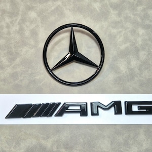 Puede incluir: Emblema negro de Mercedes-Benz con una estrella de tres puntas dentro de un marco circular. Debajo, el logotipo negro de AMG se muestra sobre un fondo rectangular blanco. El emblema y el logotipo son brillantes.