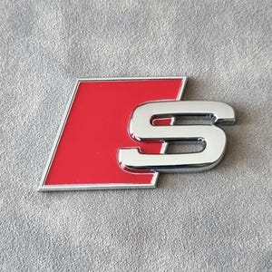 Może przedstawiać: Chromowany i czerwony emblemat z literą "S" w stylizowanym wzorze. Litera "S" ma błyszczące chromowane wykończenie, a tło jest jaskrawoczerwone. Emblemat ma srebrną obwódkę i jest umieszczony na szarym, teksturowanym tle.