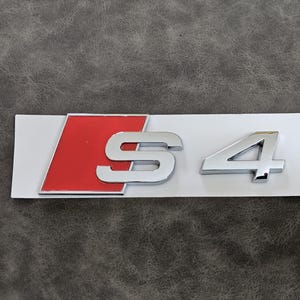May include: A chrome "S4" emblem on a white rectangular base. The "S" and "4" are in a bold, modern font. A red trapezoid shape is on the left side of the emblem, adding a striking contrast to the design.