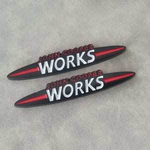 Pode incluir: Dois emblemas de carro pretos John Cooper Works. Cada emblema tem as palavras "JOHN COOPER" em vermelho e "WORKS" em branco. Uma faixa vermelha percorre o centro de cada emblema.