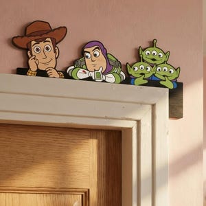 Puede incluir: Decoración de marco de puerta de madera con personajes de la película Toy Story. Los personajes incluyen a Woody, Buzz Lightyear y tres pequeños hombrecitos verdes. Las figuras están en un estilo de dibujos animados y montadas en una repisa marrón oscuro.