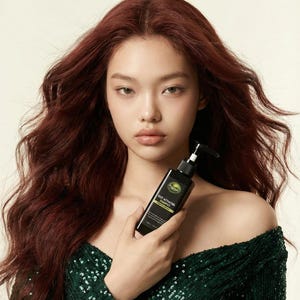 May include: A woman with long, auburn hair holds a black bottle of shampoo. The bottle has a pump dispenser and a green and white label. She wears a dark green, off-the-shoulder sequined dress. The background is a solid, light color.