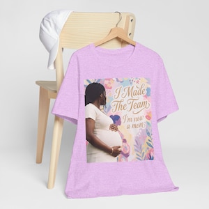 May include: A light purple t-shirt with the text "I Made The Team I'm now a mom" and a graphic of a pregnant person. The shirt is hanging on a wooden hanger. A white baseball cap rests on the back of a wooden chair.