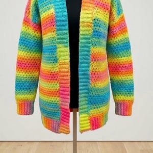 May include: A colourful, open-front crochet cardigan in rainbow hues, including pink, yellow, blue, and green. The long-sleeved sweater is displayed on a mannequin, showcasing its striped design. The garment is made of a textured yarn.