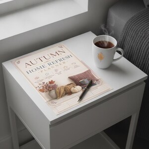Può includere: Un documento Autumn Home Refresh Planner con testo stampato e illustrazioni di fogliame autunnale, un cesto, una zucca e un cuscino. Una penna a sfera nera poggia sulla carta e una tazza in ceramica bianca con un'icona a foglia dorata si trova nelle vicinanze.
