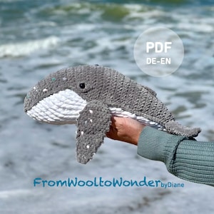 Könnte beinhalten: Ein handgefertigtes, gehäkeltes Wal-Plüschtier in Grau und Weiß mit kleinen weißen Punkten. Der Wal wird vor einem verschwommenen Ozeanhintergrund gehalten. Der Text "FromWooltoWonder byDiane" befindet sich am unteren Bildrand.