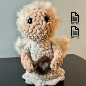 Könnte beinhalten: Handgefertigte Häkelpuppe eines Engels mit flauschigem Heiligenschein und Flügeln. Der Engel ist in einem weißen und pfirsichfarbenen Farbschema gehalten und hält ein kleines braunes Buch mit einem Herzmotiv. Die Puppe besteht aus weichem, strukturiertem Garn.