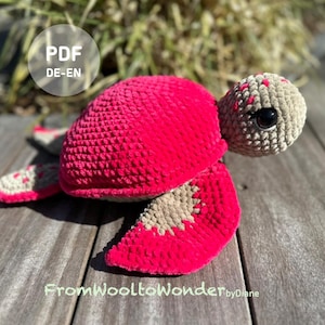 Könnte beinhalten: Eine handgefertigte Häkelschildkröte in leuchtendem Pink und Beige. Die Schildkröte hat einen strukturierten Panzer und Flossen, mit einem beigefarbenen Kopf und großen schwarzen Augen. Das Bild enthält den Text "PDF DE-EN" und "FromWooltoWonder byDiane".