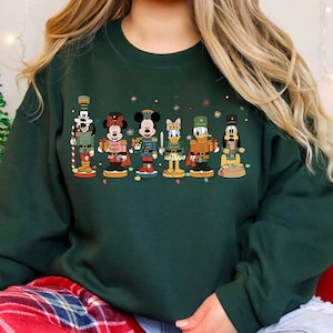 May include: A dark green crewneck sweatshirt featuring a graphic print of six Disney characters dressed as nutcrackers, including Goofy, Minnie Mouse, Mickey Mouse, Daisy Duck, Donald Duck, and Pluto, arranged in a horizontal line.