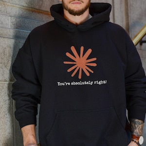 May include: A black hooded sweatshirt featuring a large, rust-colored starburst graphic centered on the chest. Below the graphic, the text "You're absolutely right!" is printed in a white, typewriter-style font. The hoodie includes a front pouch pocket.