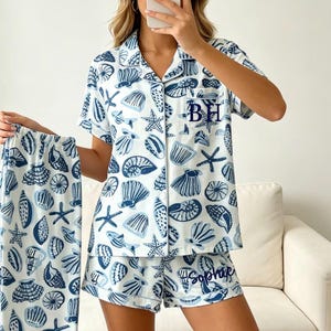 May include: A white pajama set featuring a blue seashell and starfish print. The short-sleeved button-down top has a collar, a chest pocket with the embroidered letters BH, and matching shorts with the embroidered name Sophie on the hem.