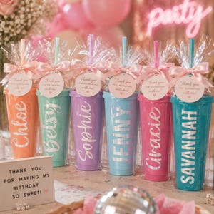 May include: Six colorful plastic tumblers with lids and straws, each wrapped in clear cellophane with a pink ribbon and a round tag reading Thank you for celebrating with me. The cups feature names in white script: Chloe, Betsy, Sophie, Jenny, Gracie, and Savannah.