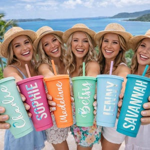 May include: Five colorful tumblers with lids and straws, each held by a person. The tumblers are in shades of teal, pink, orange, green, and light blue, each with a different name in white lettering. The people are wearing straw hats.