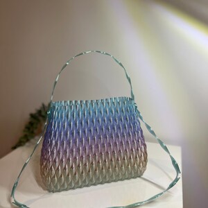 May include: A translucent handbag featuring a gradient colour transition from light blue at the top to purple in the middle and a warm beige at the base. The bag has a textured, repeating diamond-shaped pattern and a thin, matching light blue strap.