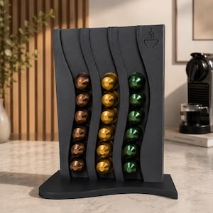 May include: A dark grey coffee pod holder with three curved sections, each holding a row of colourful coffee pods. The pods are in shades of bronze, gold, and green. A coffee cup icon is on the top right.