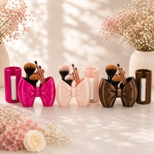 May include: Three bow-shaped makeup brush holders in hot pink, light pink, and dark brown, each paired with a matching cylindrical container holding white cotton pads. The holders feature a smooth finish and contain various makeup brushes.