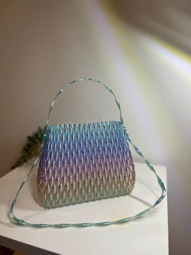 May include: A translucent handbag featuring a gradient colour transition from light blue at the top to purple in the middle and a warm beige at the base. The bag has a textured, repeating diamond-shaped pattern and a thin, matching light blue strap.