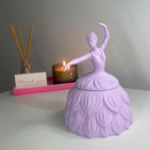 Peut inclure: Une figurine tridimensionnelle violet clair représentant une ballerine dans une pose avec un bras levé. La silhouette présente un design de jupe texturé en forme de pétales et un corsage lisse. L'objet est un récipient décoratif avec un couvercle.