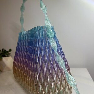 May include: A handbag featuring a gradient colour scheme transitioning from light blue at the top to purple and gold at the base. The surface has a textured, repeating geometric pattern of vertical ridges. It includes a translucent, glittery handle.