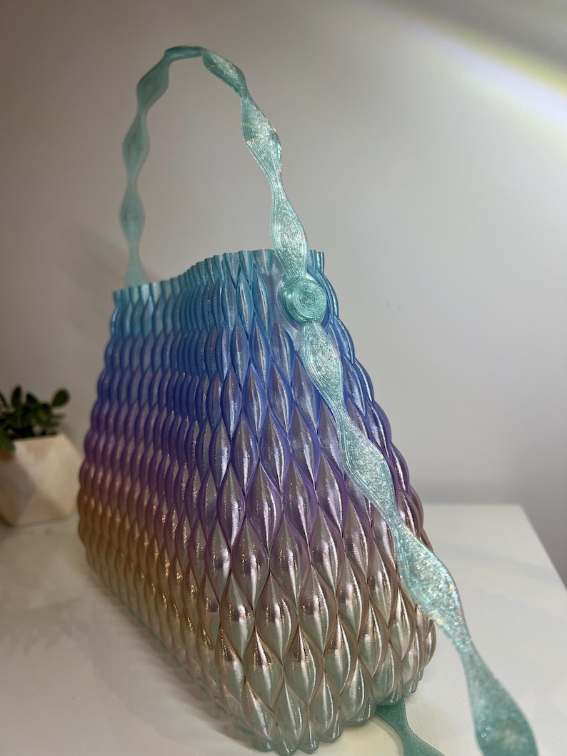 May include: A handbag featuring a gradient colour scheme transitioning from light blue at the top to purple and gold at the base. The surface has a textured, repeating geometric pattern of vertical ridges. It includes a translucent, glittery handle.