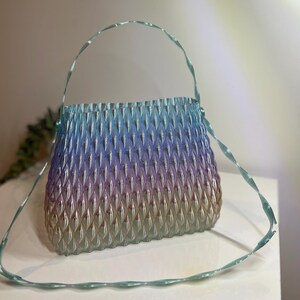 May include: A translucent handbag featuring a textured, repeating diamond-shaped pattern. The bag displays a gradient colour transition from light blue at the top, shifting through purple, to a warm beige tone at the base, with a thin, twisted handle.