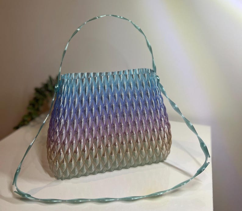 May include: A translucent handbag featuring a textured, repeating diamond-shaped pattern. The bag displays a gradient colour transition from light blue at the top, shifting through purple, to a warm beige tone at the base, with a thin, twisted handle.