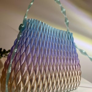 May include: A structured handbag featuring a repeating, raised leaf-like pattern across its surface. The bag displays a gradient colour transition from a light blue at the top, through purple, to a warm gold at the base. It includes a thin, translucent handle.