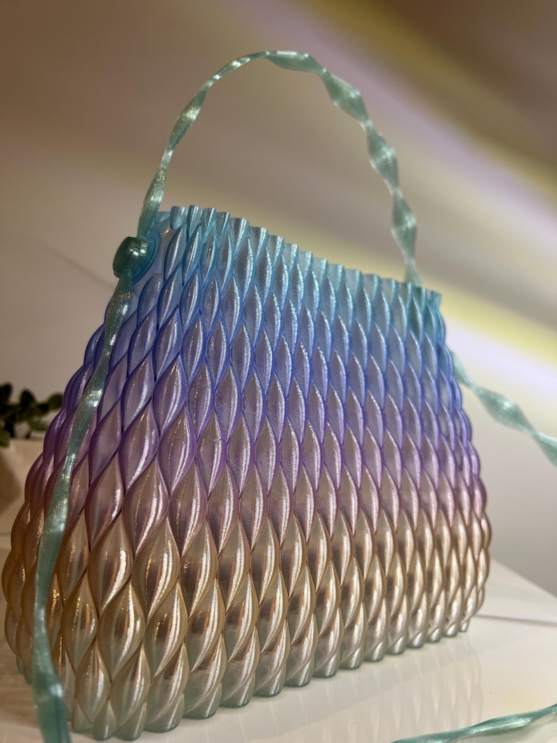 May include: A structured handbag featuring a repeating, raised leaf-like pattern across its surface. The bag displays a gradient colour transition from a light blue at the top, through purple, to a warm gold at the base. It includes a thin, translucent handle.