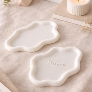 Puede incluir: Dos bandejas de cerámica blancas con forma de nube. Una bandeja tiene la palabra "Home" en relieve. Una vela encendida en un portavelas blanco acanalado y una pequeña suculenta en una maceta geométrica están en el fondo.