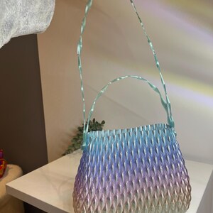 May include: A handbag featuring a gradient colour scheme that transitions from light blue at the top to purple and then to a soft gold at the base. The surface has a textured, repeating leaf-like pattern and includes a thin, twisted light blue handle.