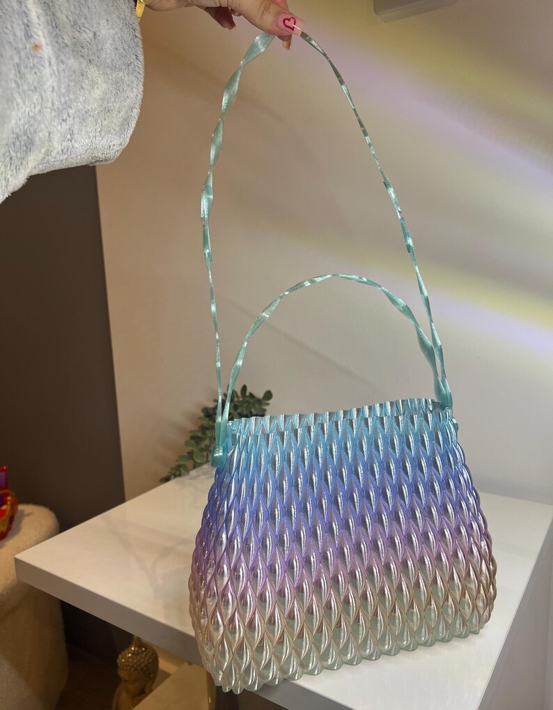 May include: A handbag featuring a gradient colour scheme that transitions from light blue at the top to purple and then to a soft gold at the base. The surface has a textured, repeating leaf-like pattern and includes a thin, twisted light blue handle.
