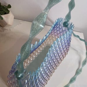 May include: A translucent, iridescent handbag featuring a repeating, scale-like textured pattern that transitions from light blue to soft purple. The bag includes a long, wavy, glittery teal handle that arches over the top of the structure.