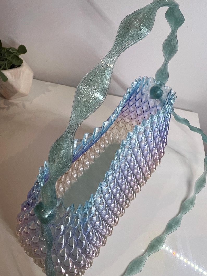 May include: A translucent, iridescent handbag featuring a repeating, scale-like textured pattern that transitions from light blue to soft purple. The bag includes a long, wavy, glittery teal handle that arches over the top of the structure.