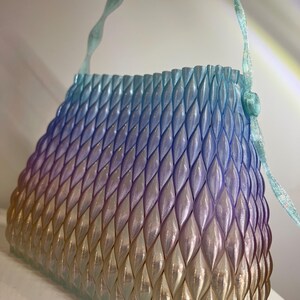 May include: A handbag featuring a structured, three-dimensional lattice pattern with a gradient colour transition from light blue at the top to purple in the middle and a warm golden yellow at the base, complete with a matching wavy handle.