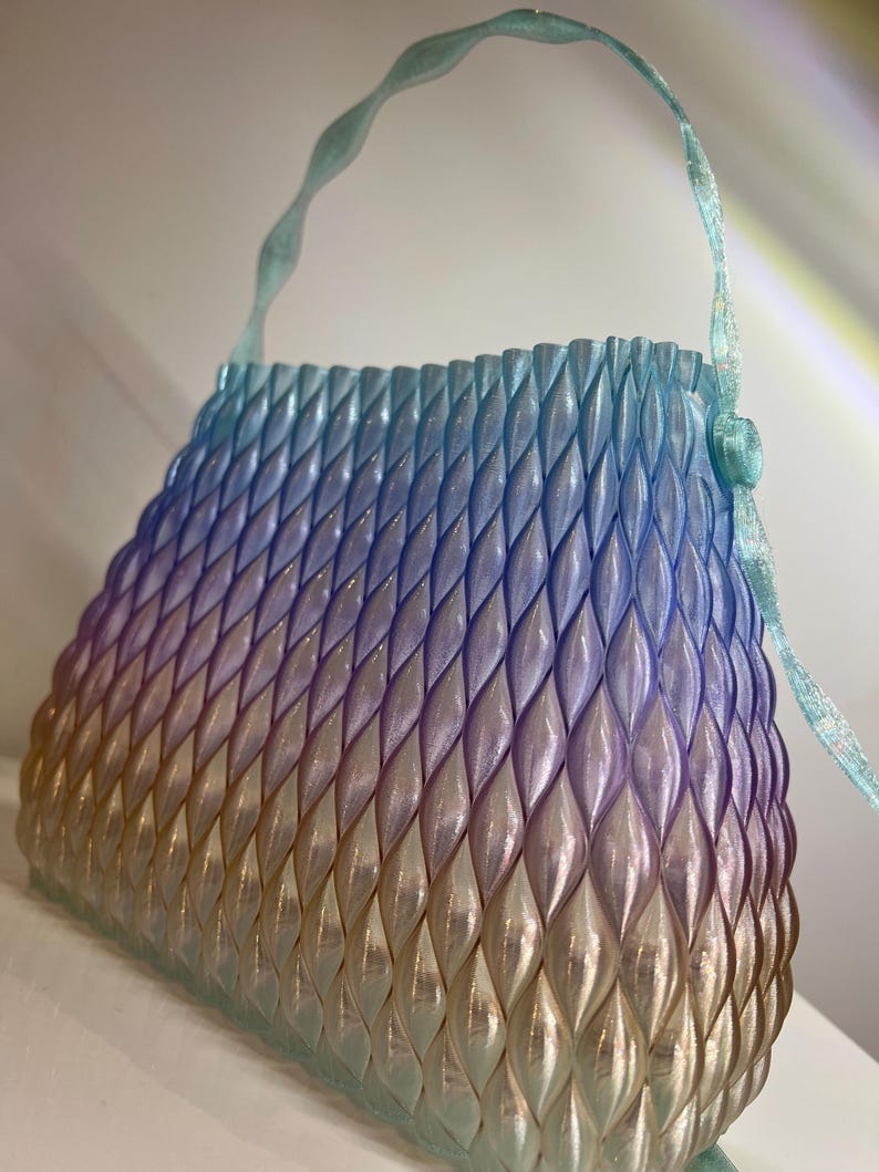 May include: A handbag featuring a structured, three-dimensional lattice pattern with a gradient colour transition from light blue at the top to purple in the middle and a warm golden yellow at the base, complete with a matching wavy handle.