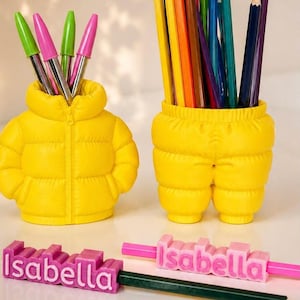 Op de afbeelding: Twee gele bureau-organizers in de vorm van een jack en een broek, gevuld met kleurrijke pennen en potloden. Twee roze rechthoekige naamblokjes met de naam Isabella in wit reliëf staan ervoor, waarvan er één een donkergroen potlood vasthoudt.
