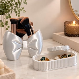 Peut inclure: Un porte-pinceaux de maquillage blanc en forme de nœud contenant plusieurs pinceaux aux poils noirs et bruns, accompagné d'un plateau ovale blanc à bords cannelés contenant divers bijoux, dont des pièces dorées avec des pierres vertes.