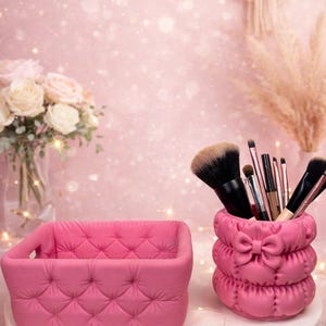May include: Pink quilted makeup brush holder and small storage basket. The brush holder has a bow detail. The basket and holder are on a white surface, with a pink background and a bouquet of roses.