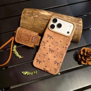 May include: A brown phone case and matching earbud case featuring a repeating pattern of Hello Kitty faces and bows. The phone case has a tan border around the camera. The earbud case has a strap. The cases are on a dark surface.