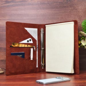 May include: A brown faux leather portfolio folder with a three-ring binder mechanism, featuring multiple interior pockets, a zippered compartment, a white pen, and a grid-lined notepad. A silver smartphone lies on the surface in front of the open folder.
