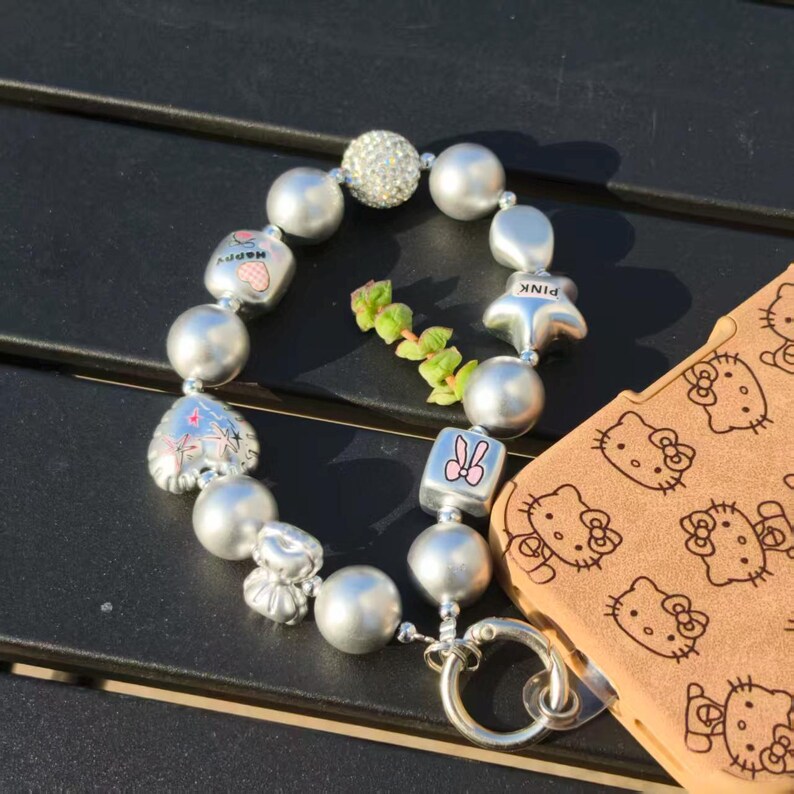 May include: A silver beaded bracelet with charms, including a heart, star, and a cube with a bunny design. The bracelet is next to a phone case with a repeating Hello Kitty pattern.