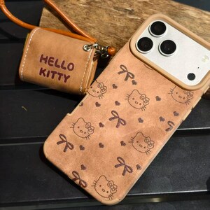 May include: A brown phone case and matching small pouch with a strap. The phone case features a pattern of Hello Kitty faces, bows, and hearts. The pouch has the words "HELLO KITTY" printed on it.