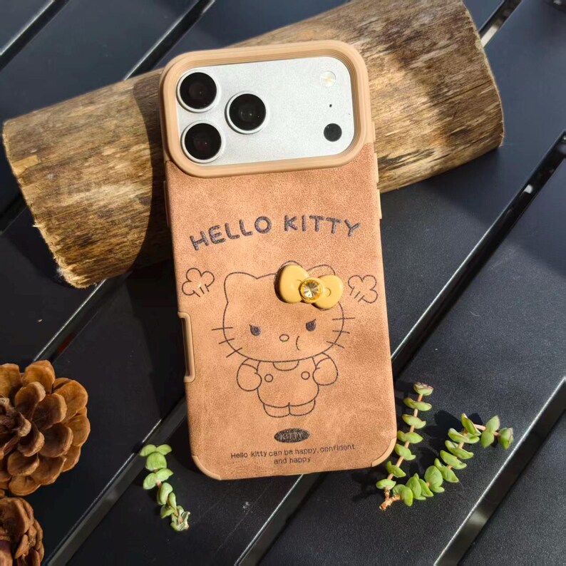 May include: Brown phone case featuring a Hello Kitty design. The case has a tan border around the camera and a yellow bow with a gem. The words "HELLO KITTY" are embossed on the case, along with the phrase "Hello kitty can be happy, confident and happy."
