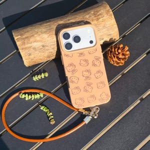 May include: A tan smartphone case featuring a repeating pattern of Hello Kitty character outlines. The case includes a camera cutout and a detachable orange wrist strap with a silver metal clasp, providing a secure way to carry the device.